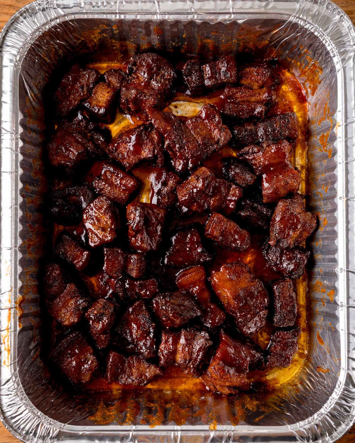 Cubed pieces of cooked, glazed pork belly arranged in a disposable foil tray, each coated with a rich caramelized sauce - Pork Belly Burnt Ends.