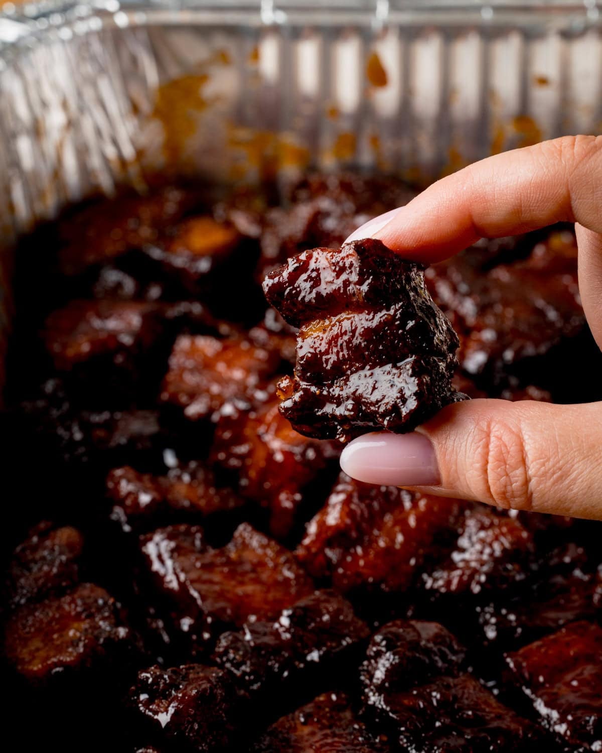 A hand has picked up and is squeezing a a piece of glazed Pork Belly Burnt End - above a tray filled more pork belly bites.