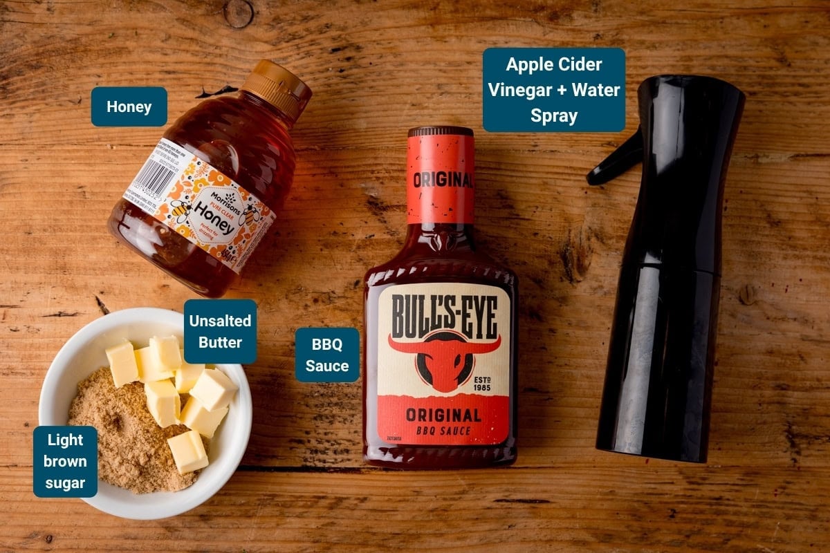 A bottle of honey, a bowl with light brown sugar and unsalted butter, a bottle of BBQ sauce, and a spray bottle labelled apple cider vinegar + water on a wooden table. All ingredients are labelled - for the glaze for pork belly bites.