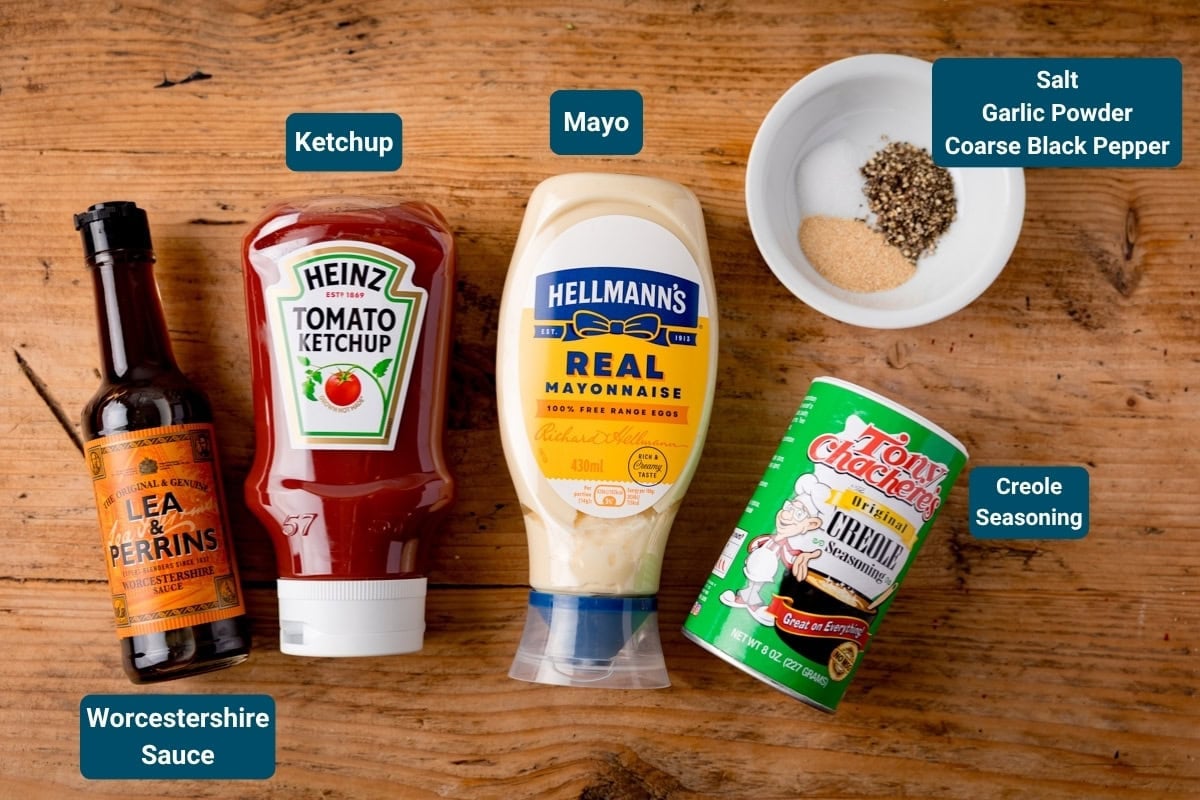 Bottles of Worcestershire sauce, tomato sauce, and mayonnaise, a tin of Creole seasoning, and a bowl with salt, garlic granules, and coarse black pepper on a wooden surface&mdash;perfect for making a Cane's sauce recipe copycat.