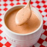 A spoon holds a portion of creamy, orange-coloured copycat Cane's sauce with black pepper specks above a white ramekin. The ramequin sits on a red and white chequered surface.
