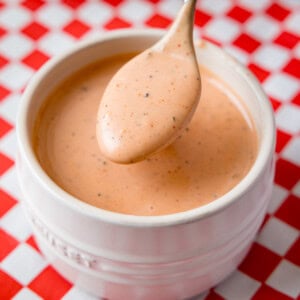 A spoon holds a portion of creamy, orange-coloured copycat Cane's sauce with black pepper specks above a white ramekin. The ramequin sits on a red and white chequered surface.