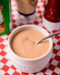 A white dish filled with creamy copycat Cane's sauce. A spoon is sitting in the sauce. The dish is on a red and white chequered surface, with condiment bottles in the background.