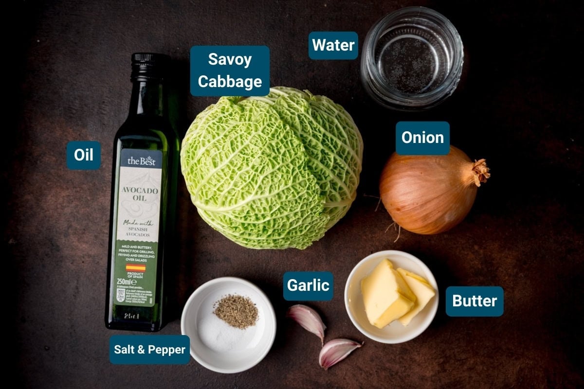 Ingredients for sauteed cabbage on a dark background, including: A head of savoy cabbage, an onion, garlic cloves, butter, avocado oil, water, and a small dish of salt and pepper.