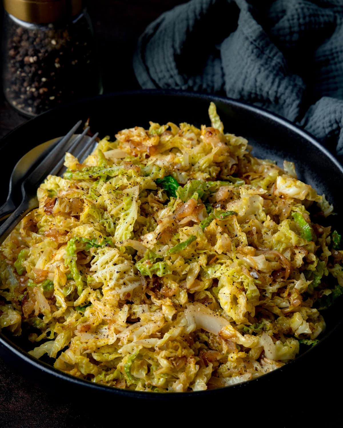 Side-on image of a black bowl filled with sautéed cabbage, next to a fork, knife, peppermill and dark napkin.