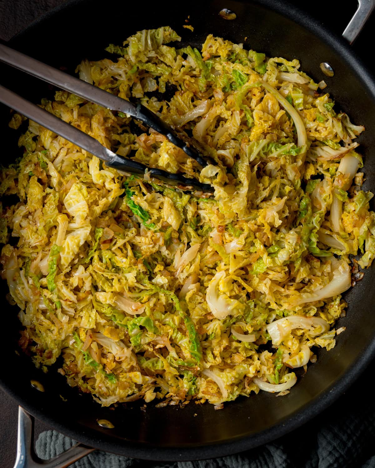 Overhead image of a pan filled with sautéed cabbage and onions, with metal tongs resting on top.