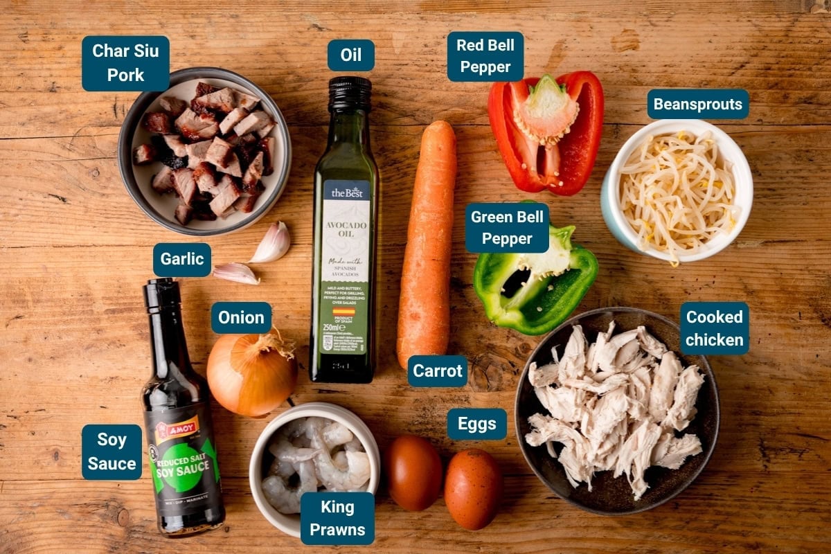A wooden table displaying labelled ingredients for a Singapore Noodle stir fry: char siu pork, cooked chicken, king prawns, eggs, veg, soy sauce, garlic, onion and oil.