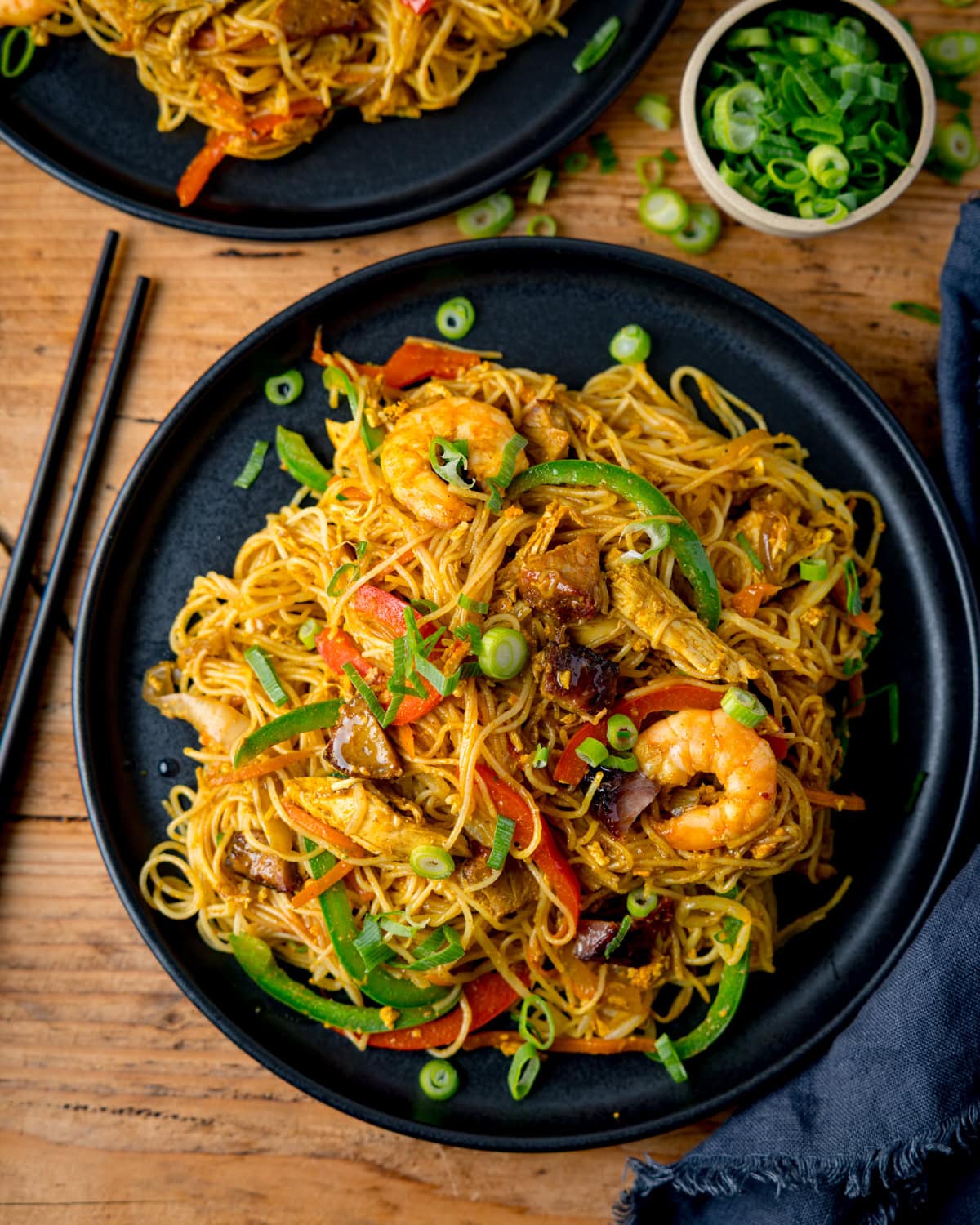 A black plate of stir-fried Singapore noodles with prawns, chicken, vegetables, and spring onions on a wooden table with chopsticks and a small bowl of sliced spring onions nearby.
