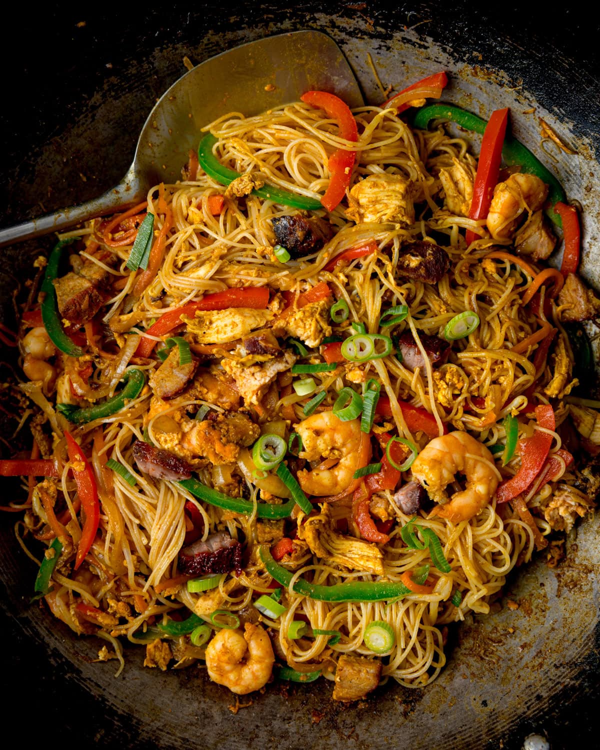 A wok filled with stir-fried rice noodles, prawns, chicken, pork, sliced vegetables, spring onions, with a large metal spatula on the side.