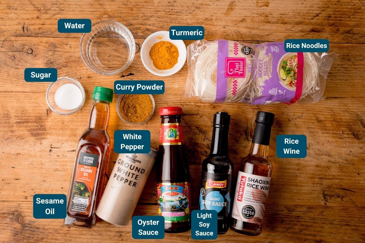 A variety of labelled cooking ingredients for Singapore Noodles sauce and noodles on a wooden surface, including rice noodles, sauces, spices, water, and sugar.
