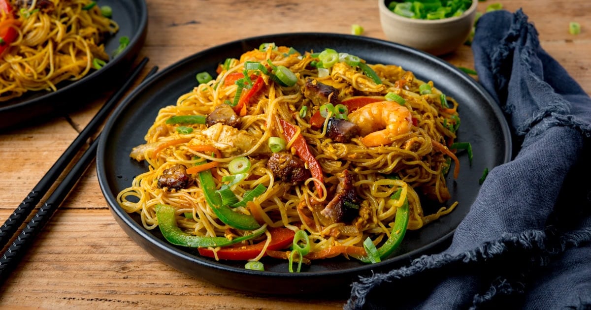 A black bowl with thin noodles, sliced peppers, prawns, crispy pieces of pork and sliced chicken.