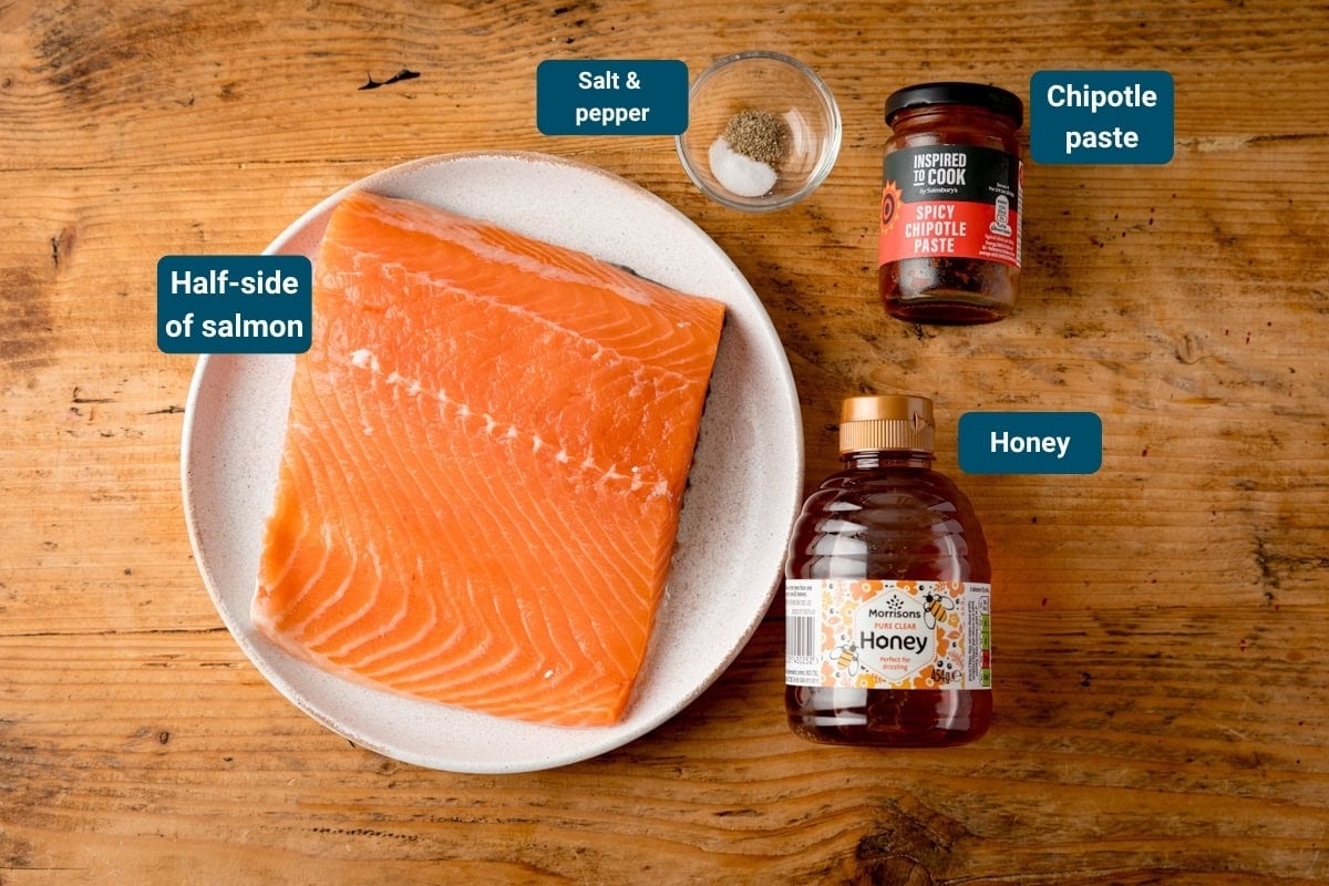 A half-side of raw salmon on a plate, with small bowls of salt and pepper, a jar of chipotle paste, and a bottle of honey on a wooden surface.