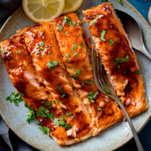 Glazed salmon fillets garnished with chopped herbs on a light blue plate, with lemon slices and a fork at the side.