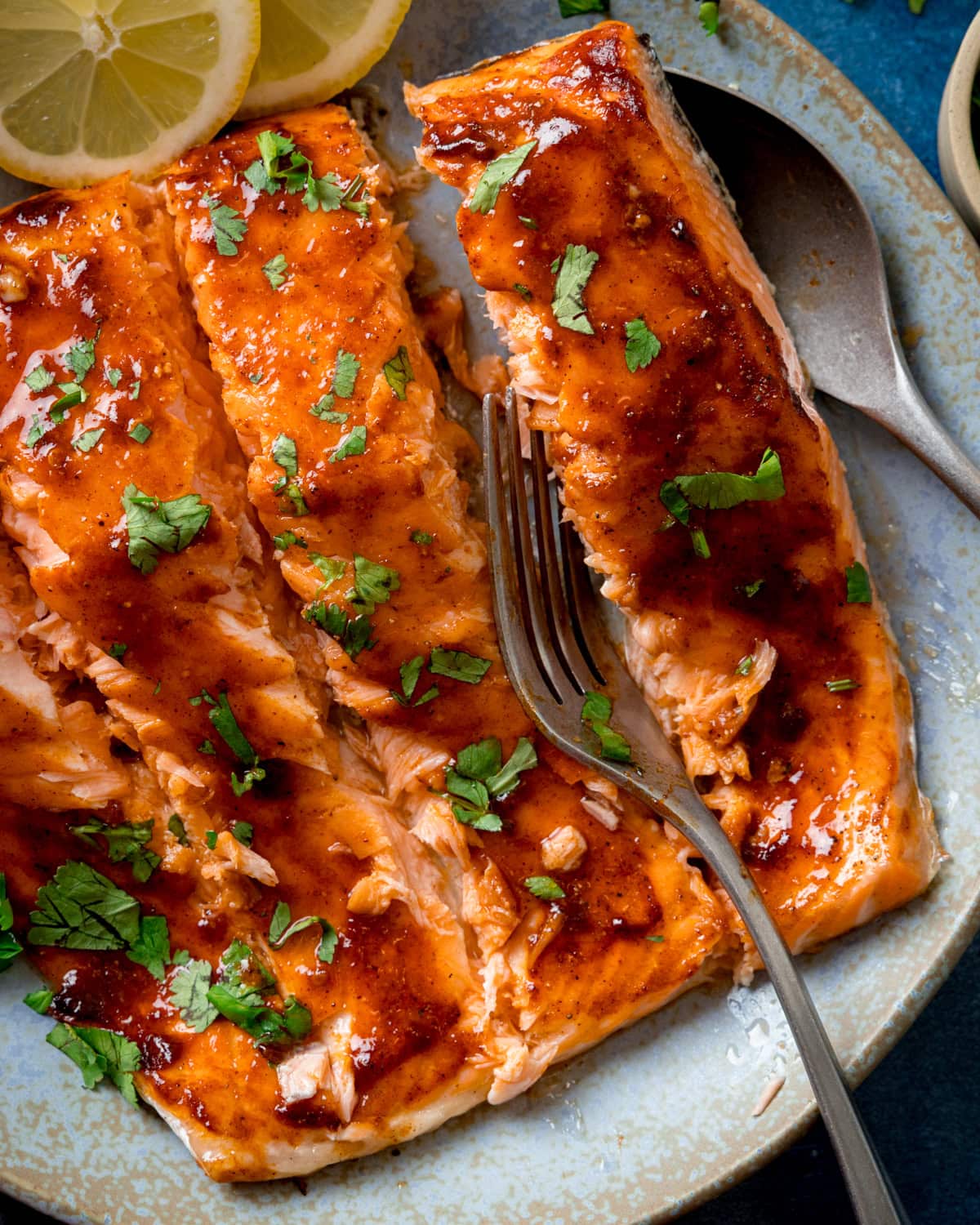 Glazed side of salmon sliced into fillets and garnished with chopped parsley on a light blue plate, with a fork flaking one fillet and lemon slices on the side.