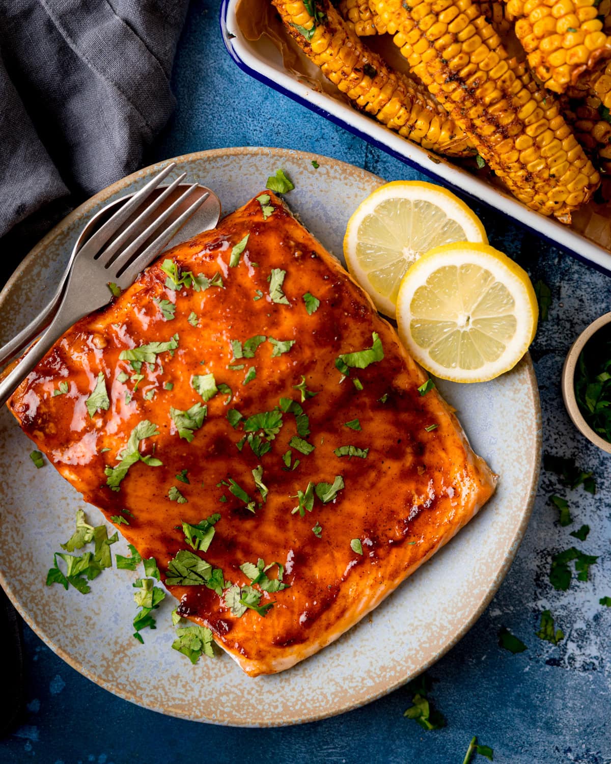 A glazed salmon fillet topped with chopped parsley on a plate, with two lemon slices and cutlery, next to a tray of grilled corn ribs.