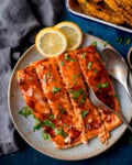 Glazed side of salmon sliced into fillets and garnished with chopped parsley on a light blue plate, with lemon slices on the side and a fork and spoon beside the fish.