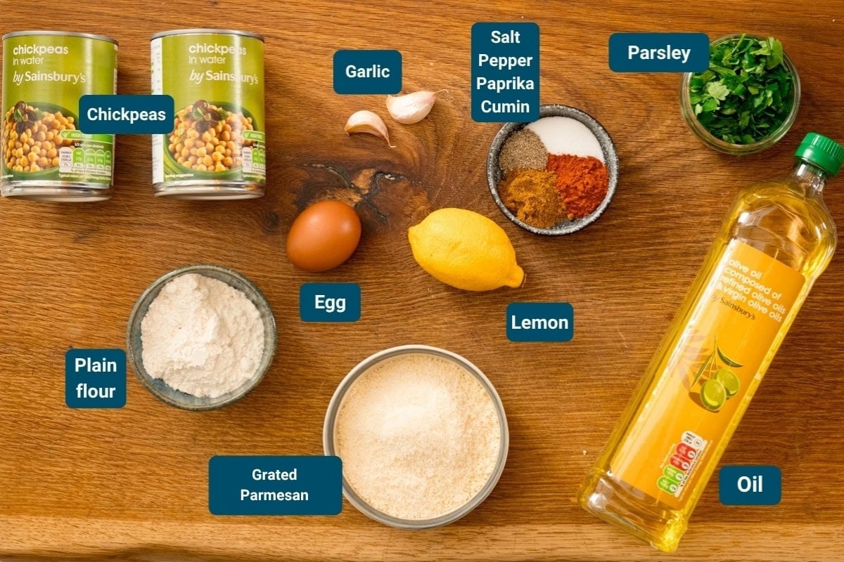 Ingredients for making chickpea fritters on a wooden surface: tinned chickpeas, garlic, spices, parsley, plain flour, egg, lemon, panko breadcrumbs, and oil.