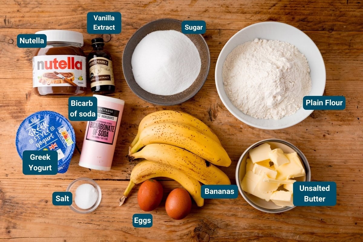 Ingredients on a wooden surface for Nutella Banana Bread including Nutella, vanilla extract, sugar, plain flour, butter, bananas, eggs, Greek yoghurt, bicarbonate of soda, and salt—all ingredients are labelled.