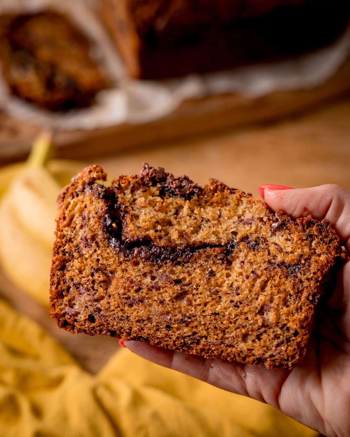 A hand holds a slice of Nutella Banana Bread with a chocolate swirl, with the remaining loaf and a banana in the background on a wooden surface.