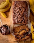 Top-down view of a loaf of Nutella-swirled banana bread on a wooden board, with two sliced pieces, bananas, and a bowl of Nutella next to it.