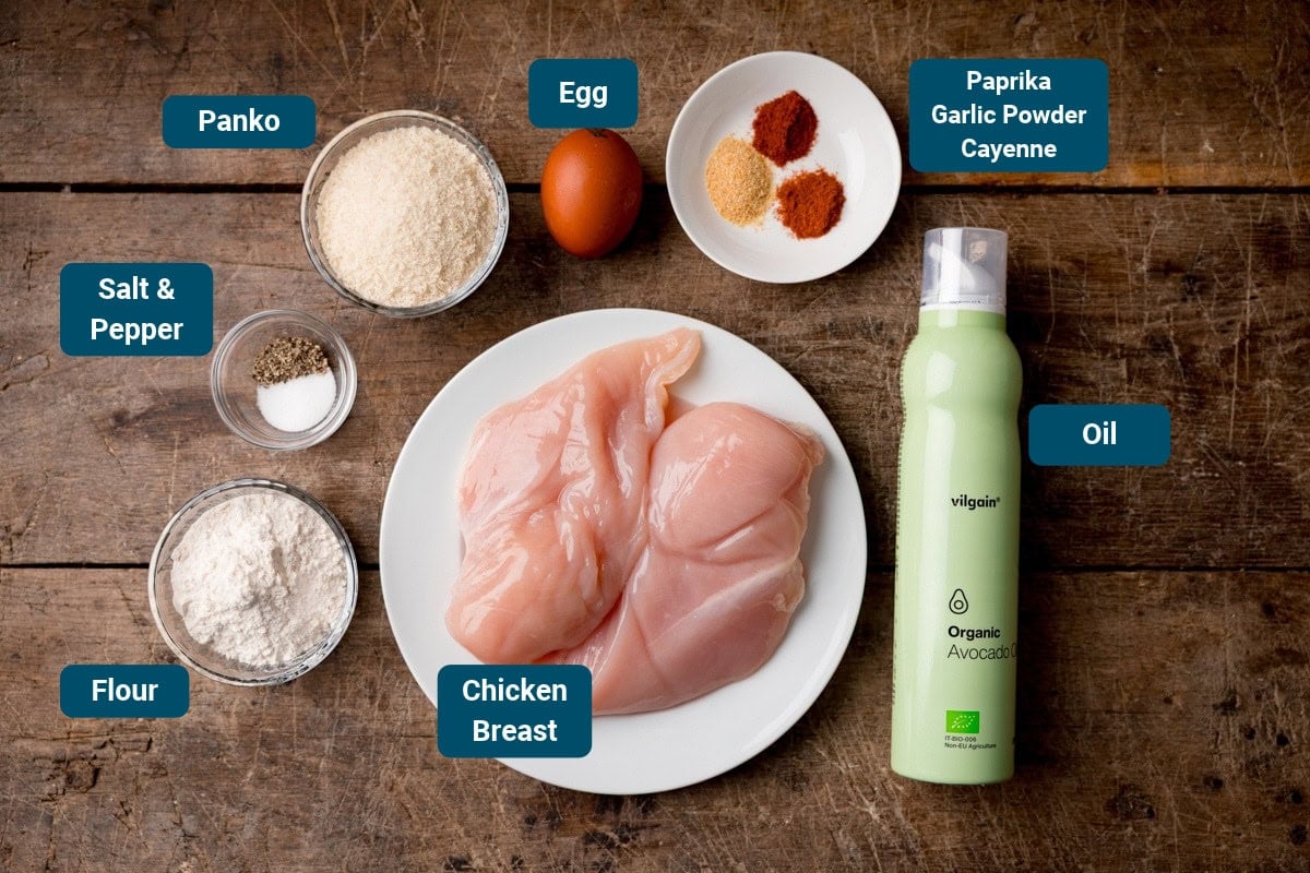 Ingredients for crispy chicken, including chicken breast, flour, egg, panko, oil spray, salt, pepper, paprika, garlic powder, and cayenne, labelled on a wooden surface.