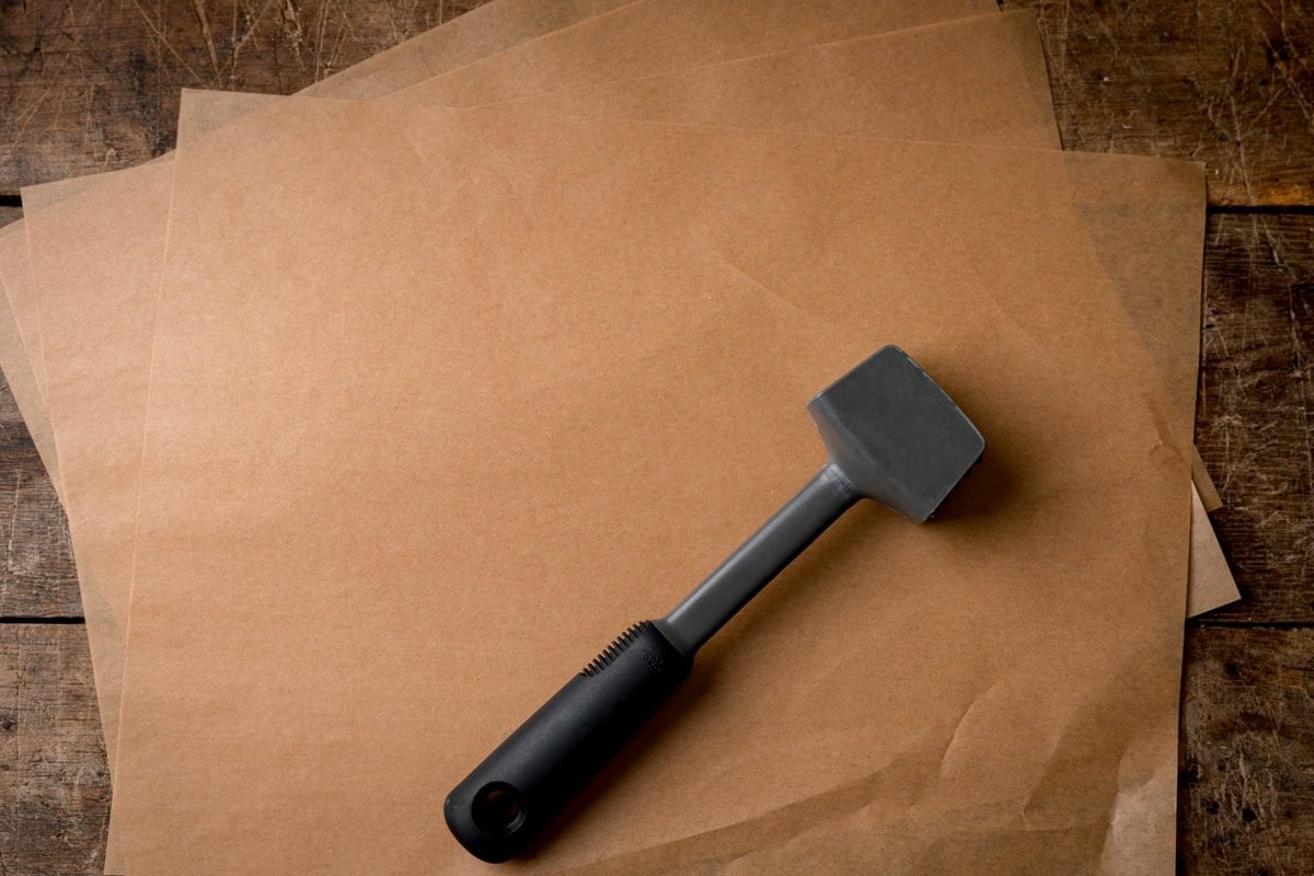 A grey meat mallet for preparing rests on sheets of greaseproof paper on a wooden surface.