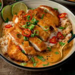A bowl of crispy sliced chicken served over rice with a creamy curry sauce, garnished with chillies, spring onions, and coriander, served with a lime wedge.