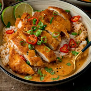 A bowl of crispy sliced chicken served over rice with a creamy curry sauce, garnished with chillies, spring onions, and coriander, served with a lime wedge.