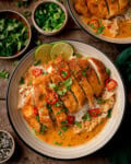 Sliced crispy chicken breast on brothy rice in a creamy coconut sauce, garnished with chilli, spring onion, coriander, and lime wedges.