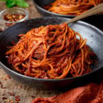 A black bowl filled with Spaghetti all’Assassina in a red tomato sauce, with a small dish of red chilli flakes in the background.