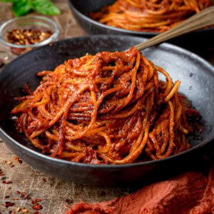 A black bowl filled with Spaghetti all’Assassina in a red tomato sauce, with a small dish of red chilli flakes in the background.