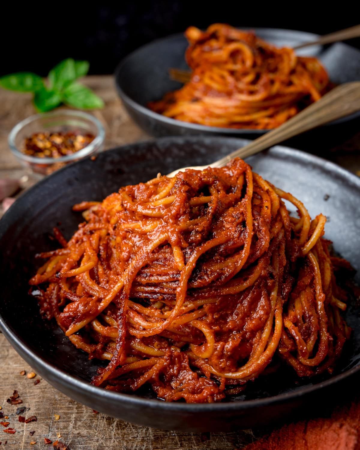 A black bowl filled with Spaghetti all’Assassina in red tomato sauce, with a second portion in the background and a small dish of red chilli flakes.