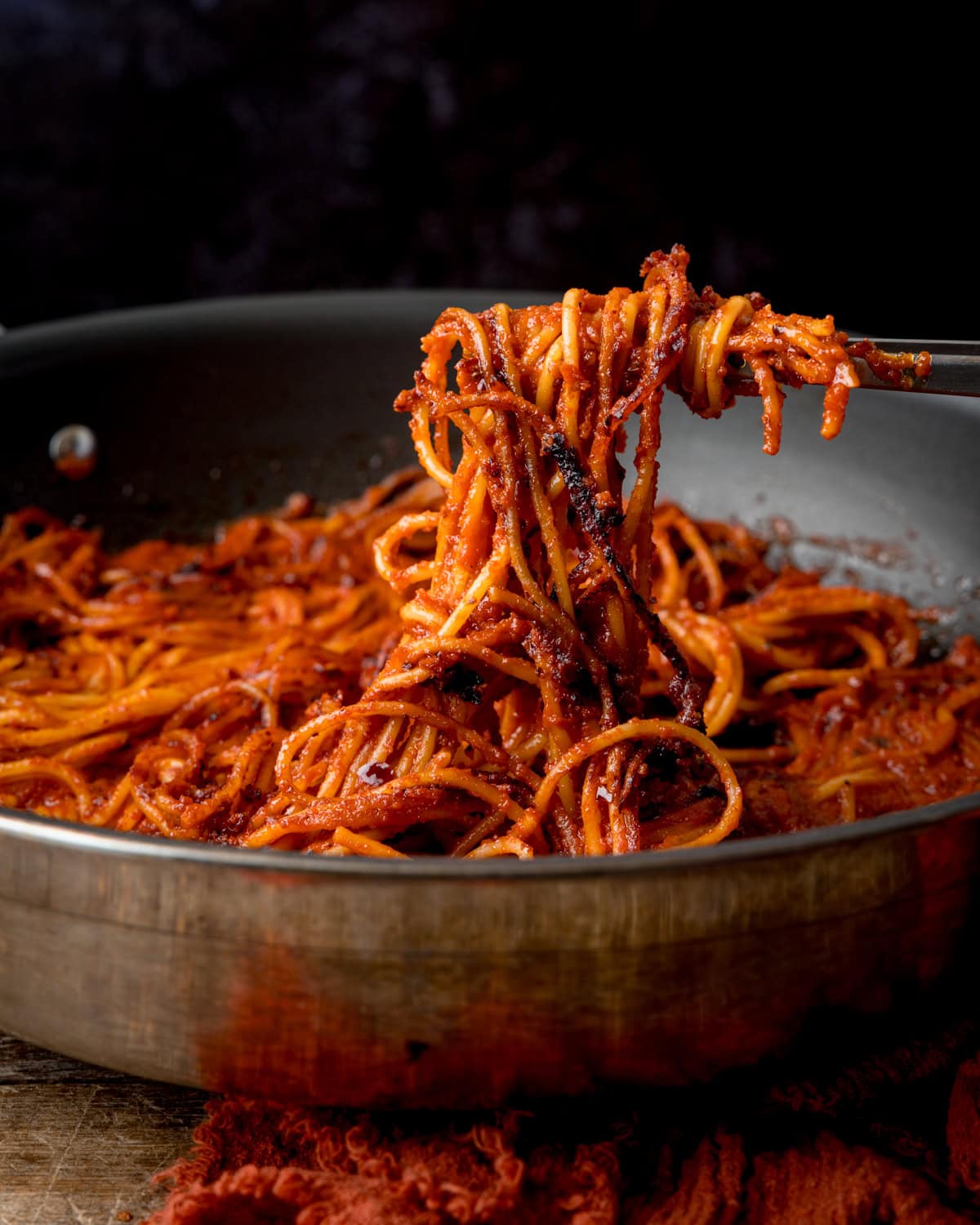 A fork lifting a portion of Spaghetti all’Assassina coated in red tomato sauce from a saucepan.