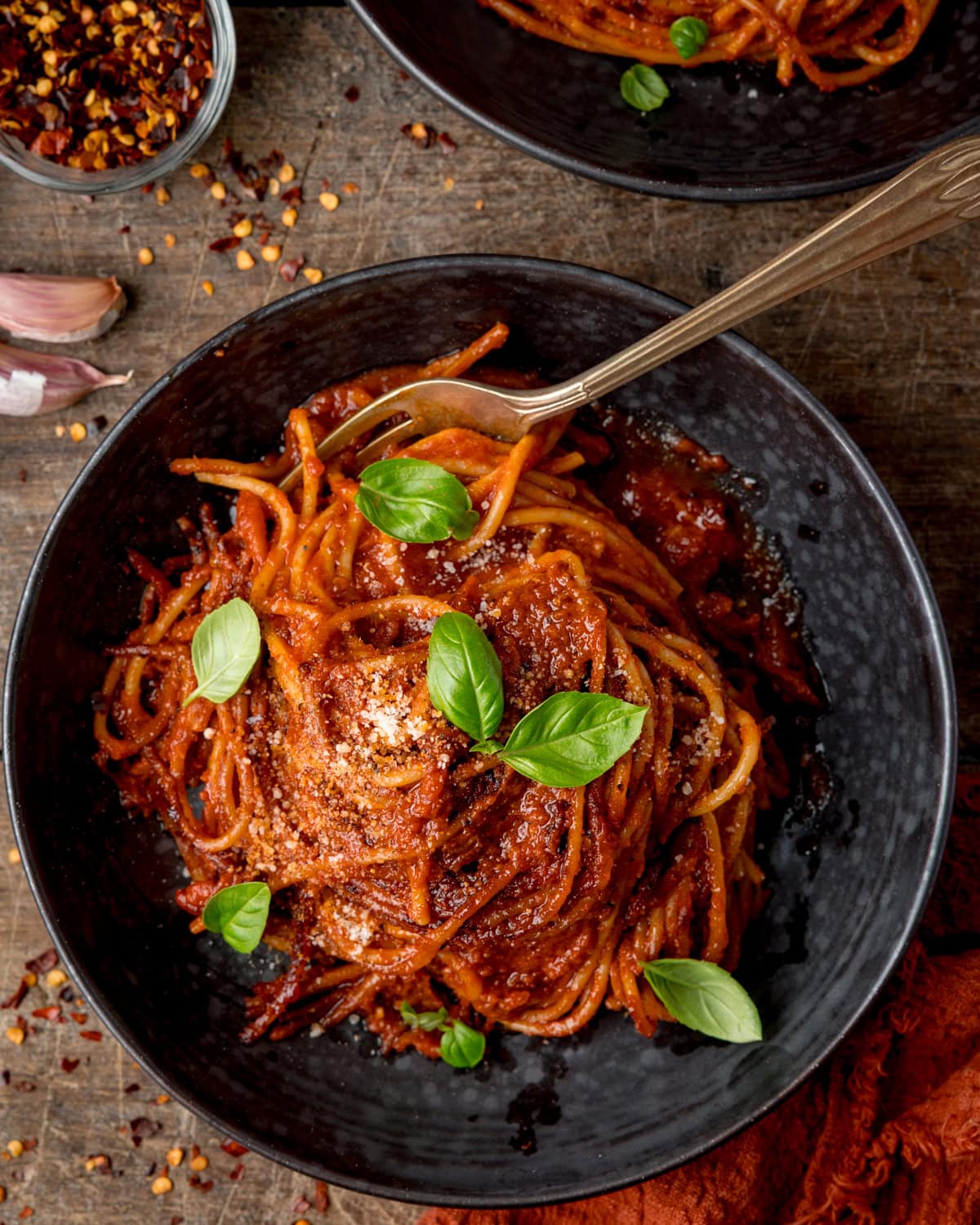 A dark bowl of spicy tomato spaghetti sprinkled with parmesan and fresh basil leaves, with a fork resting inside.
