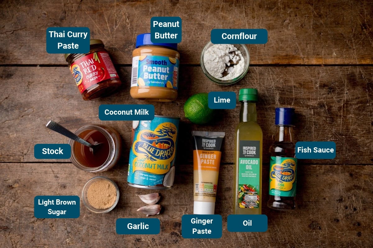 Ingredients for Thai peanut-coconut sauce arranged on a wooden surface: Thai curry paste, peanut butter, cornflour, stock, coconut milk, lime, garlic, ginger paste, avocado oil, soft brown sugar, and fish sauce.