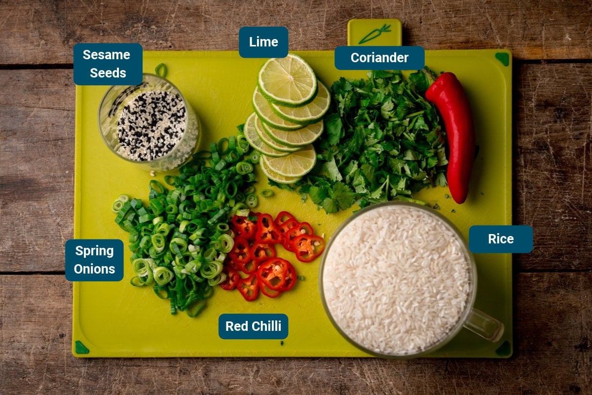 Green chopping board on a wooden surface, with labelled ingredients for brothy rice toppings, including sesame seeds, lime, coriander, spring onions, red chilli, and the rice.