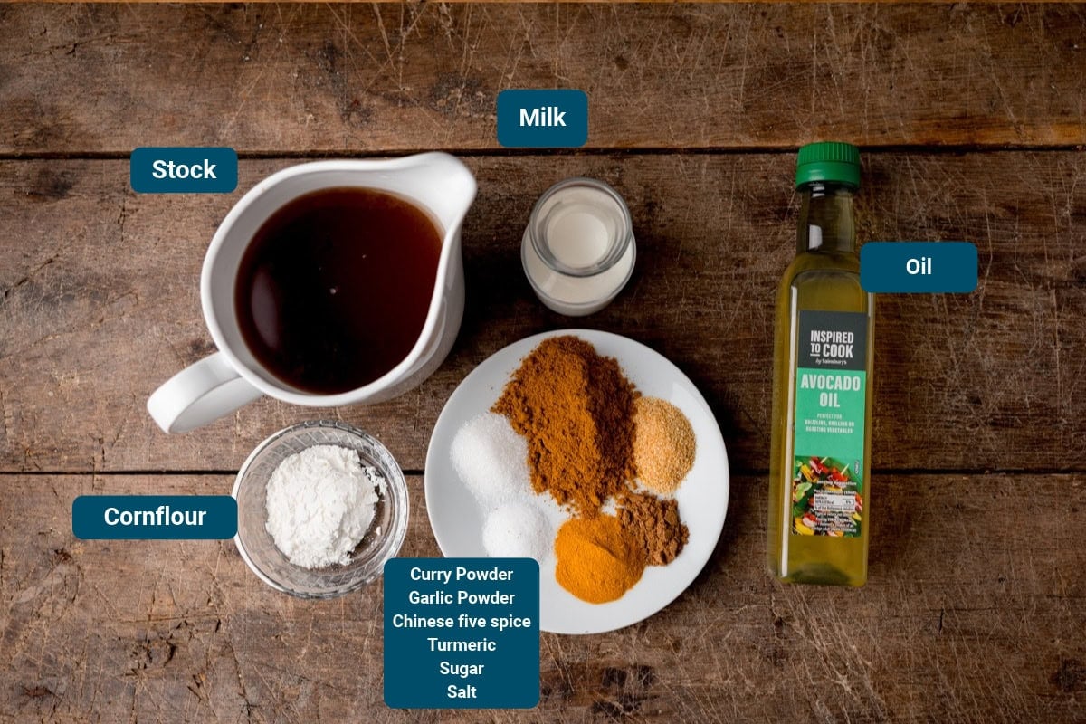 A jug of stock, milk, avocado oil, and bowls of cornflour and spices for Chinese curry sauce, including curry powder, garlic, five spice, turmeric, sugar, and salt on a wooden table. All ingredients are labelled.