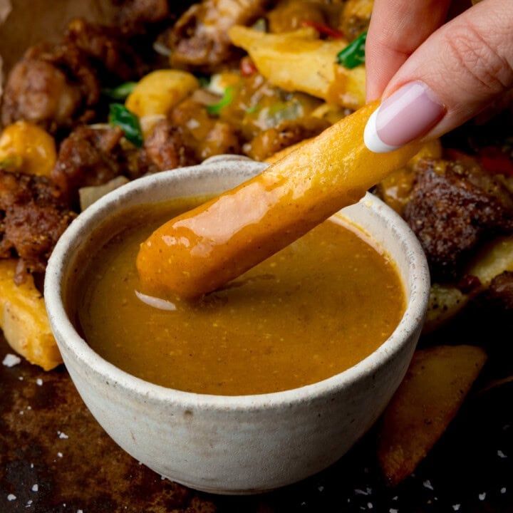 A chip being dipped into a bowl of Chinese style curry sauce with a portion of salt and pepper chicken in the background.