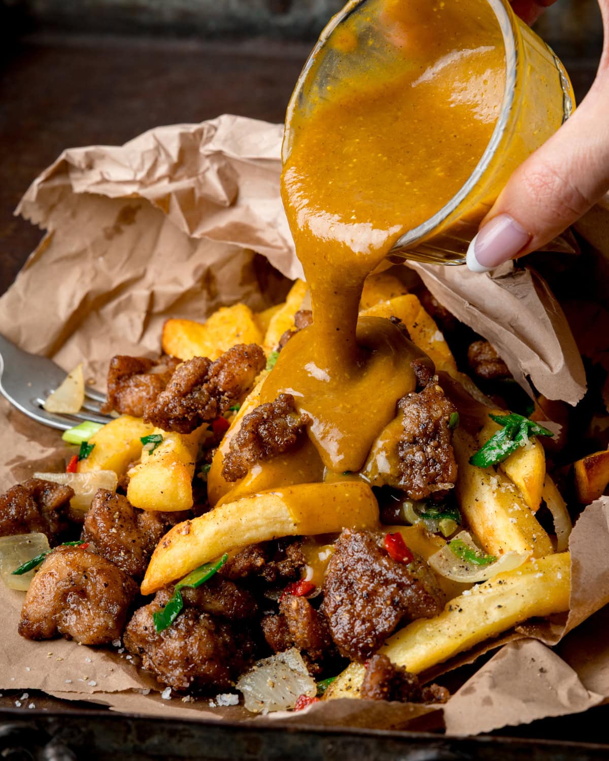 Chinese curry sauce being poured from a glass dish over crispy chicken and chips and fried meat in a paper-lined bag.