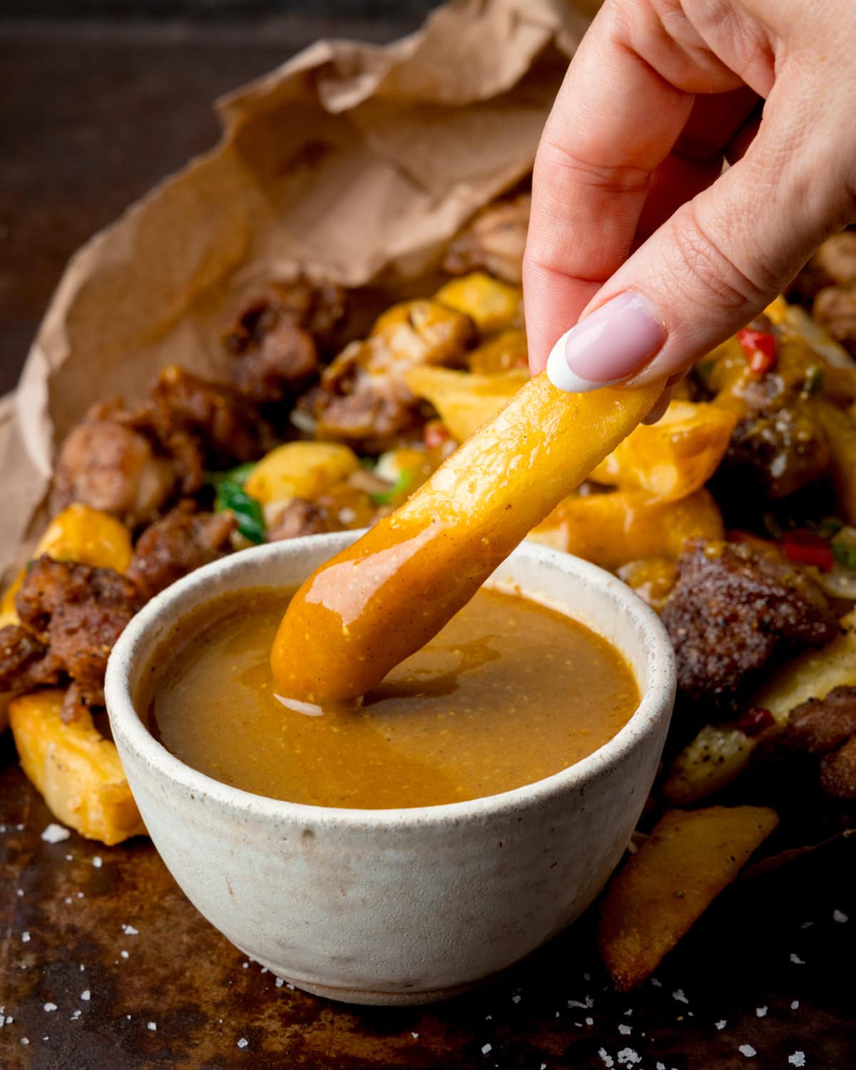 Tall image of a hand dipping a chip into a small bowl of Chinese curry sauce, with a portion of salt and pepper chips in the background.