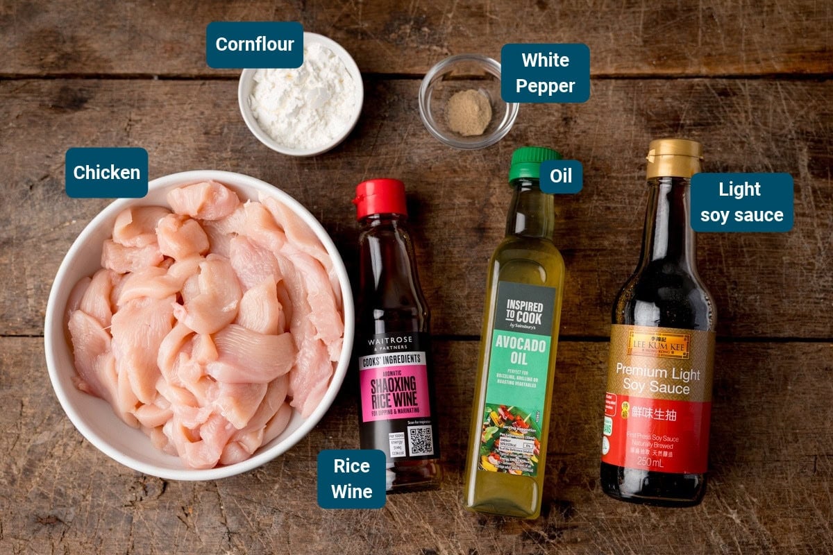 Ingredients for Hunan chicken including sliced chicken, a small bowl of cornflour, white pepper, and bottles of rice wine, avocado oil, and light soy sauce arranged on a wooden surface.