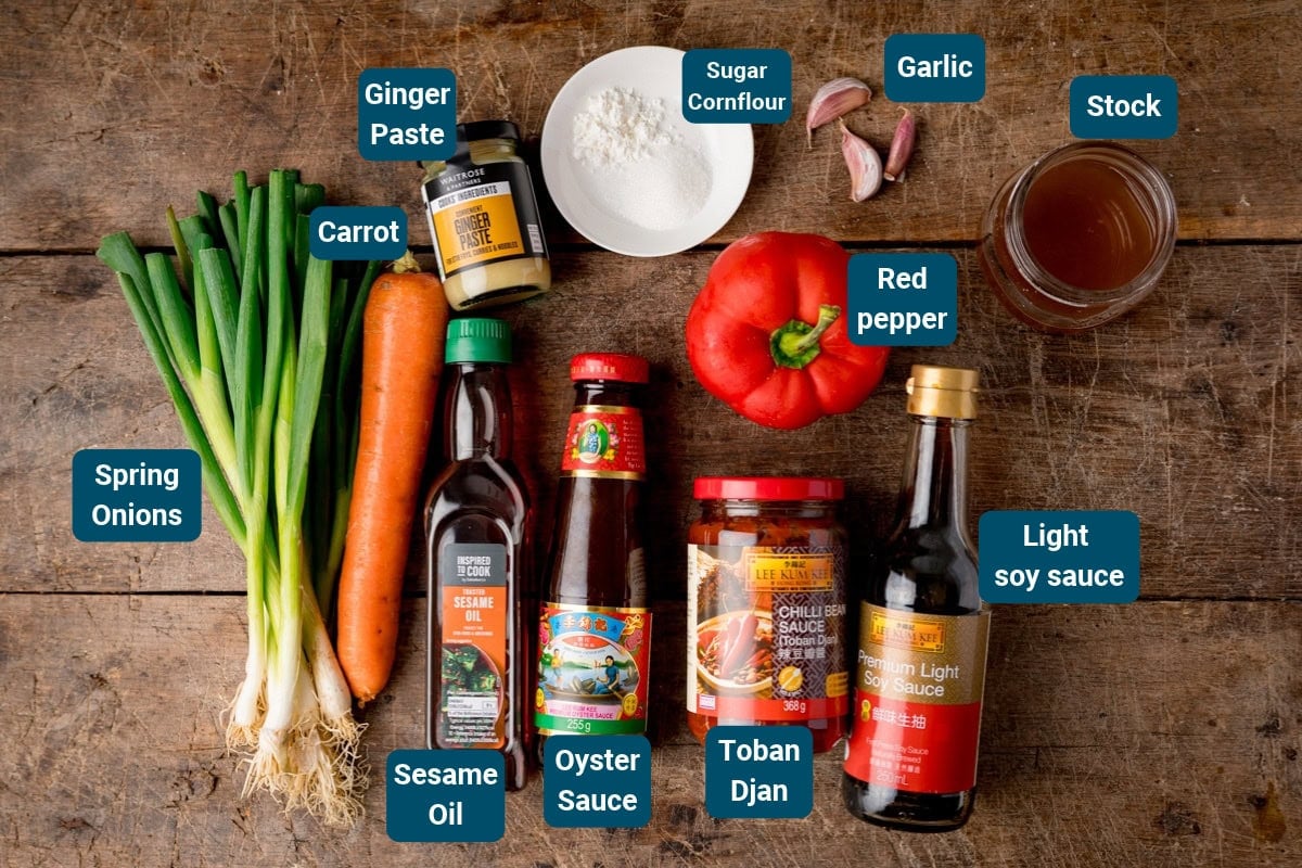 Ingredients for the sauce for Hunan Chicken on a wooden surface, including spring onions, carrot, ginger paste, cornflour, garlic, red pepper, stock, sesame oil, oyster sauce, toban djan, and light soy sauce.
