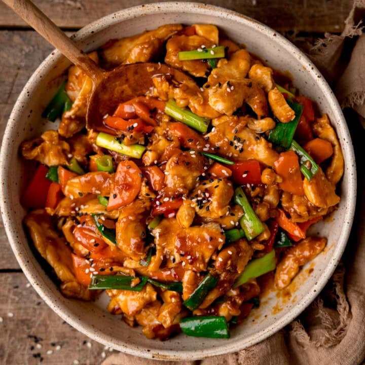 Spicy Hunan chicken stir-fried with peppers and spring onions in a bowl with a wooden spoon, topped with sesame seeds.