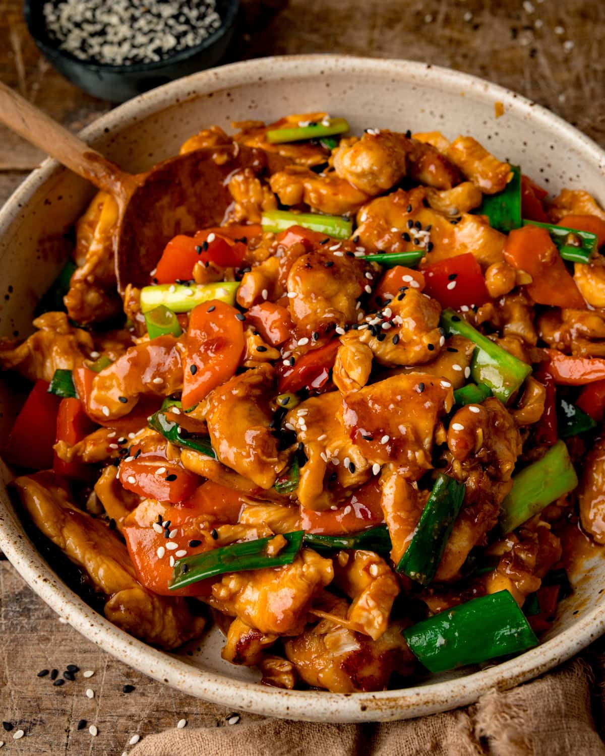 A bowl of Hunan-style chicken stir-fry with peppers and spring onions, garnished with sesame seeds and served with a wooden spoon.