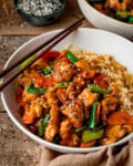 A bowl of spicy Hunan chicken stir-fry with vegetables and rice, topped with sesame seeds and served with chopsticks in a white bowl.