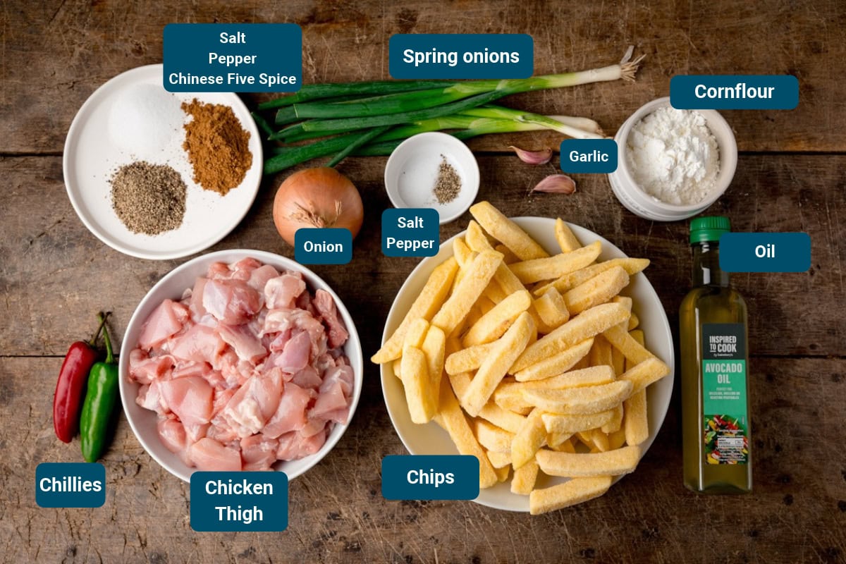 Ingredients for an Irish spice bag, including chicken thigh pieces, chips, spring onions, onion, garlic, oil, chillies, and various seasonings. Ingredients are on a wooden table, all labelled.