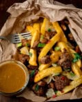 A takeaway spice bag consisting of salt and pepper chips, chicken, onions, and spring onions in a paper bag, served with a side of curry sauce and a fork.