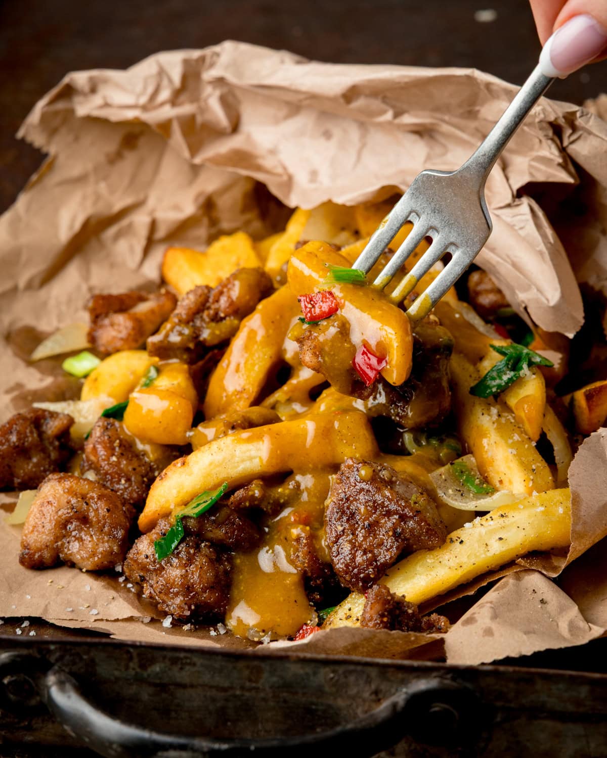 A fork lifts chips from an Irish spice bag, topped with curry sauce, pieces of salt and pepper chicken, and spring onions from a paper-lined tray.