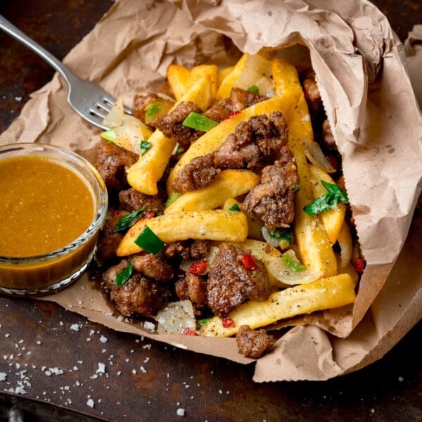 An Irish spice bag consisting of salt and pepper chips, chicken, onions, and chillies served in brown paper with a side of curry sauce and a fork.