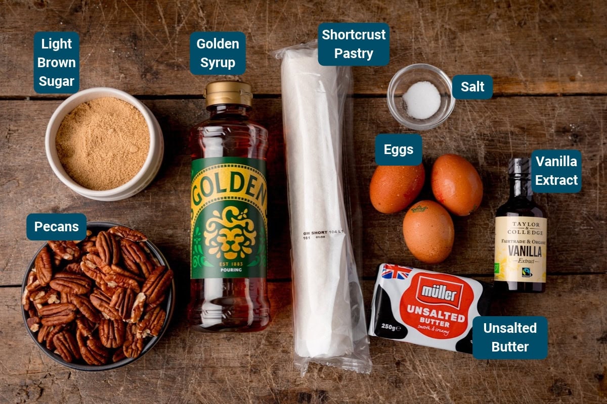Ingredients for a pecan pie on a wooden table: light brown sugar, golden syrup, shortcrust pastry, salt, eggs, vanilla extract, and unsalted butter.